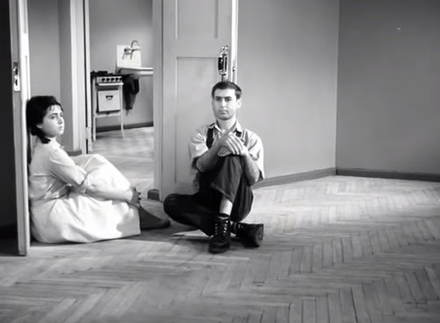 Black and white still from the film April, the young couple sit on the floor of an otherwise empty apartment.